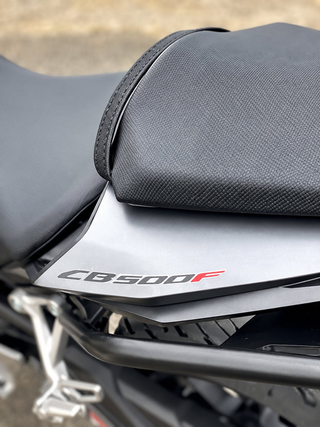 Gros plan sur la selle passager et le carénage arrière gris d'une moto portant l'inscription CB500F en lettres rouges et grises.