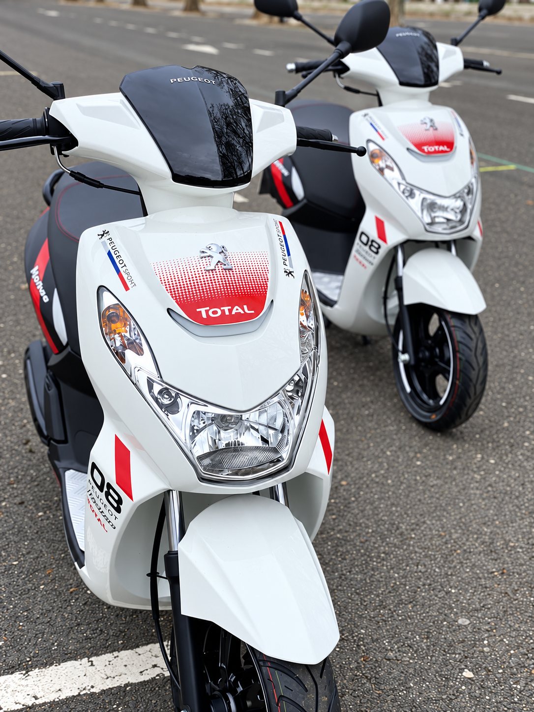 Deux scooters Peugeot blancs ornés de décorations Peugeot Sport et Total, garés côte à côte sur un parking en bitume.