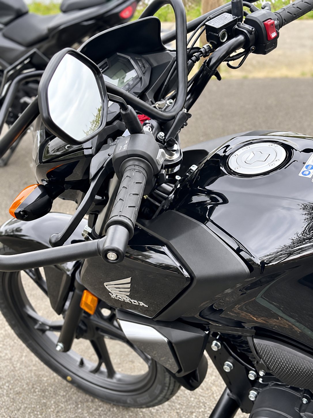 Gros plan sur le guidon, le rétroviseur et le réservoir d'une moto Honda noire garée à l'extérieur, avec une autre moto floue à l'arrière-plan.