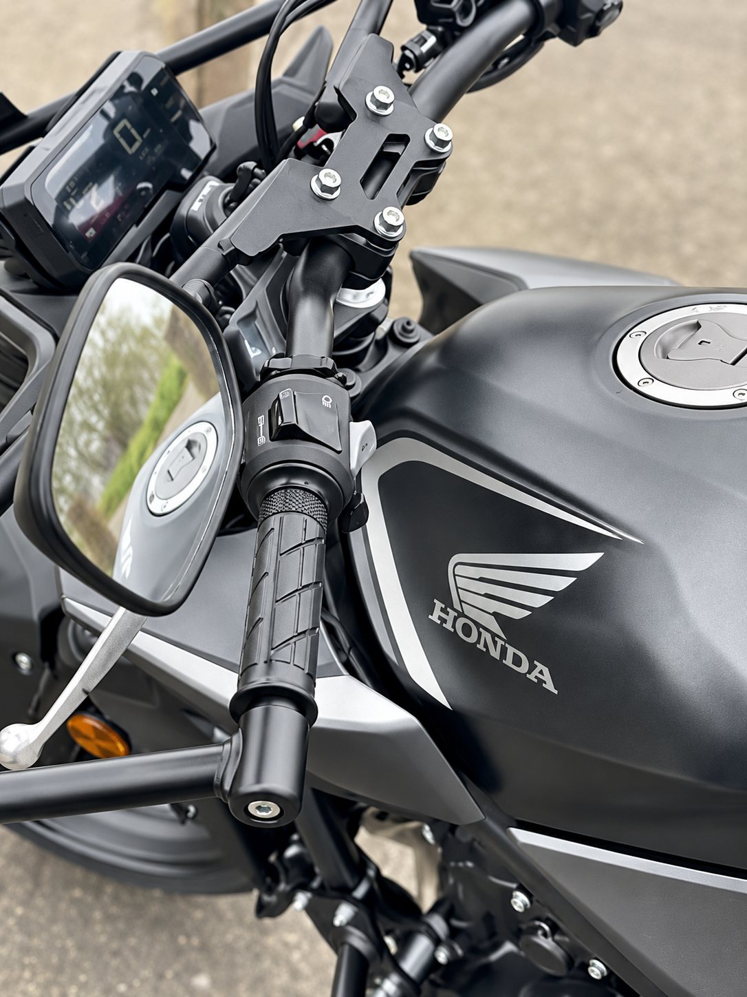 Vue rapprochée du guidon, du rétroviseur et du réservoir noir mat orné du logo Honda d'une moto garée à l'extérieur.