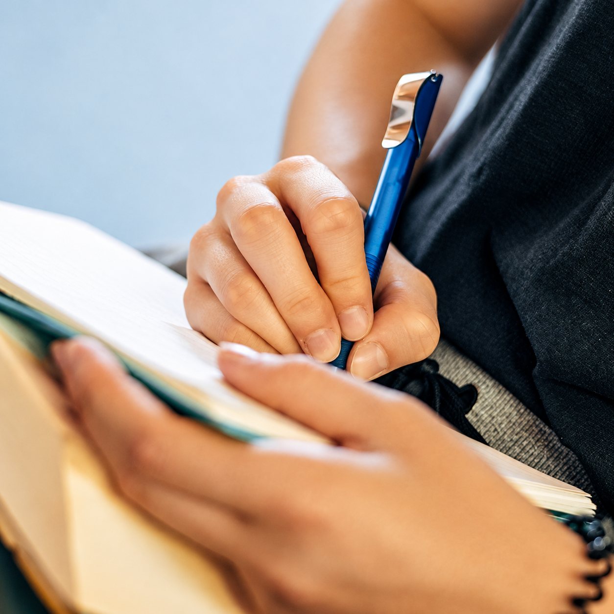 Gros plan sur les mains d'une personne qui écrit avec un stylo bleu dans un carnet ouvert posé sur ses genoux.