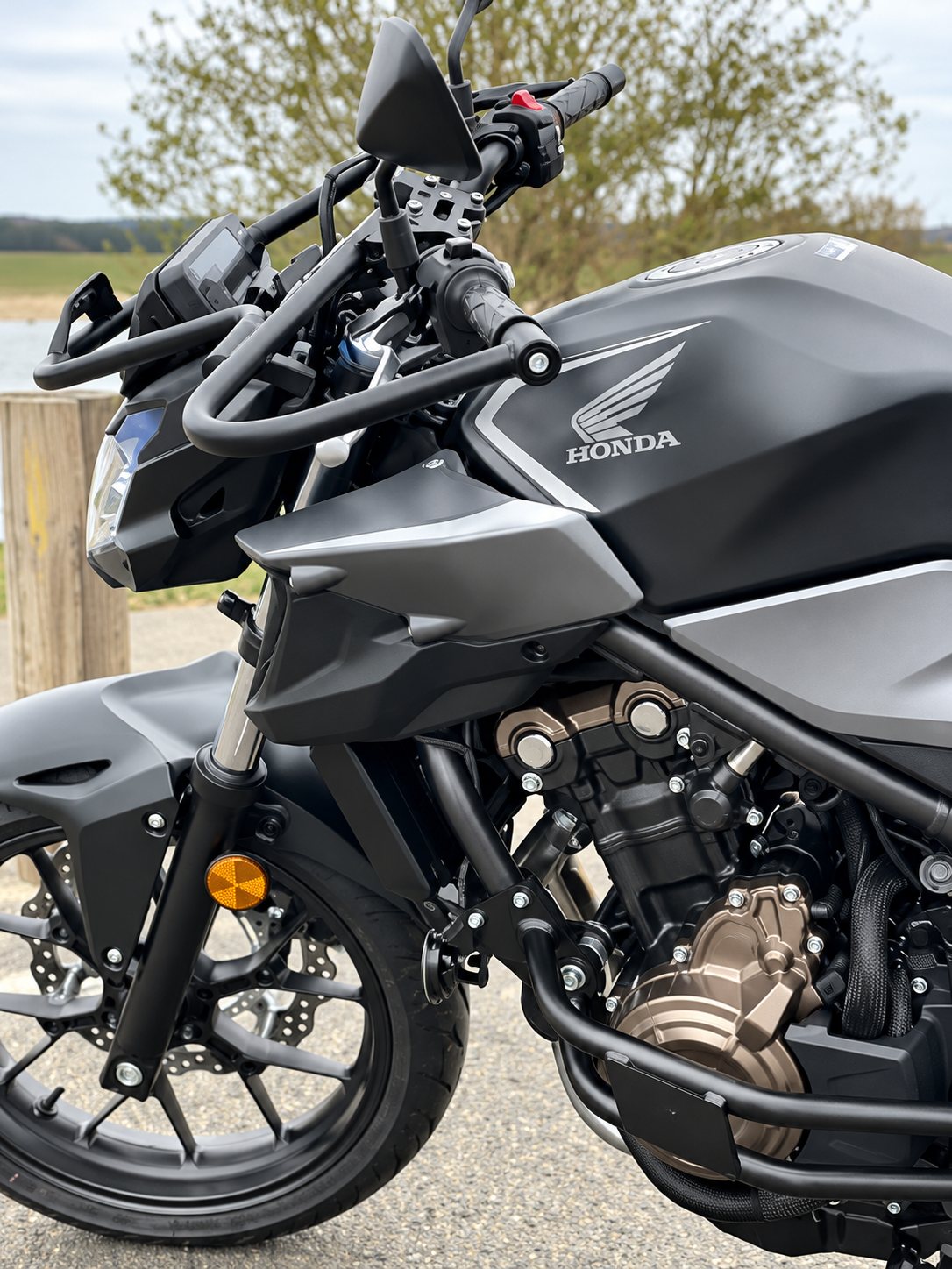 Gros plan sur le côté d'une moto Honda noire mate, avec son moteur, son guidon et sa roue avant, garée à l'extérieur.