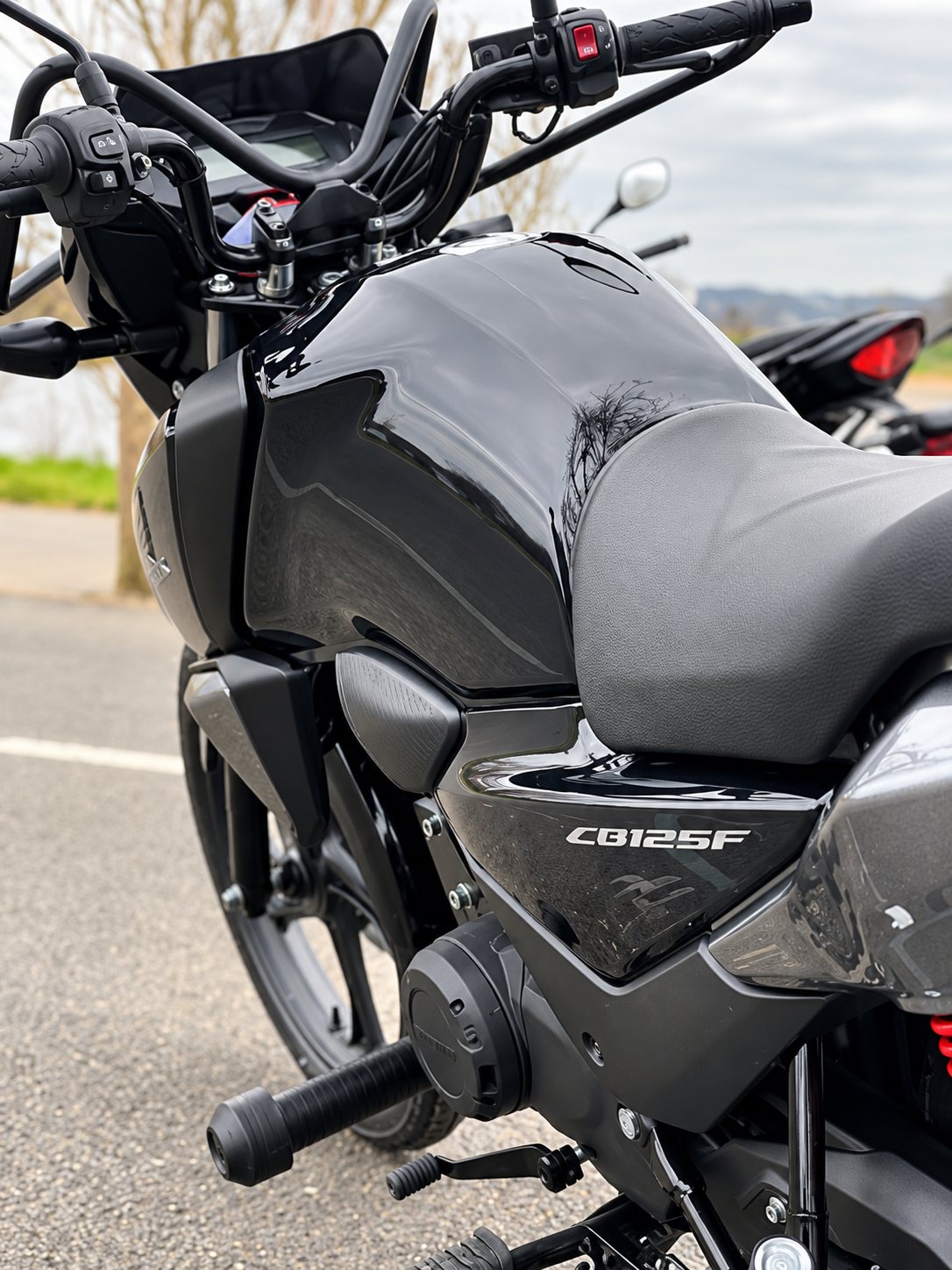 Gros plan sur une moto noire de modèle CB125F, avec son réservoir, sa selle et son guidon, garée sur un parking à l'extérieur.