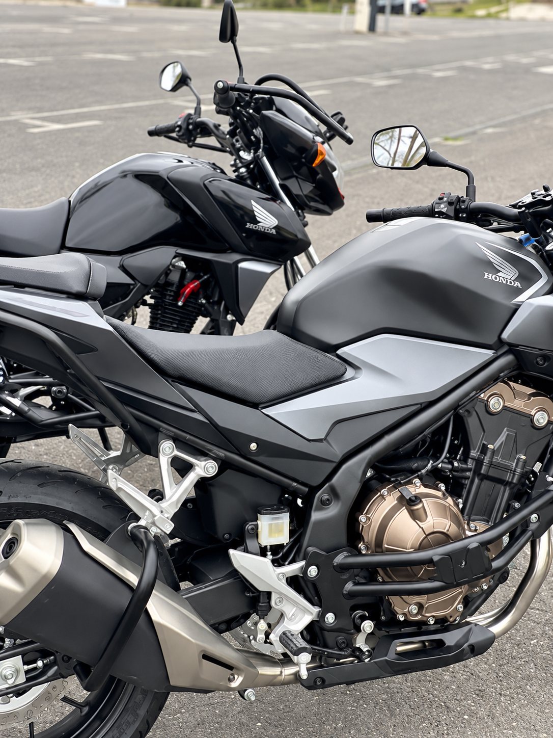 Deux motos Honda noires stationnées côte à côte sur un parking, l'une au premier plan avec son moteur et son échappement visibles.
