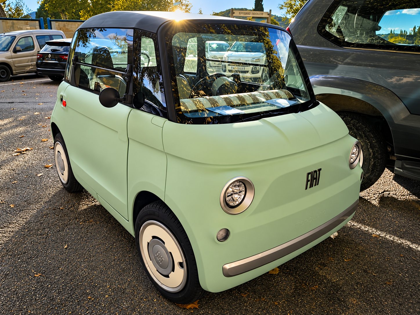 Une petite voiture électrique Fiat vert pastel est garée sur un parking, entre deux autres véhicules, sous un soleil rasant.