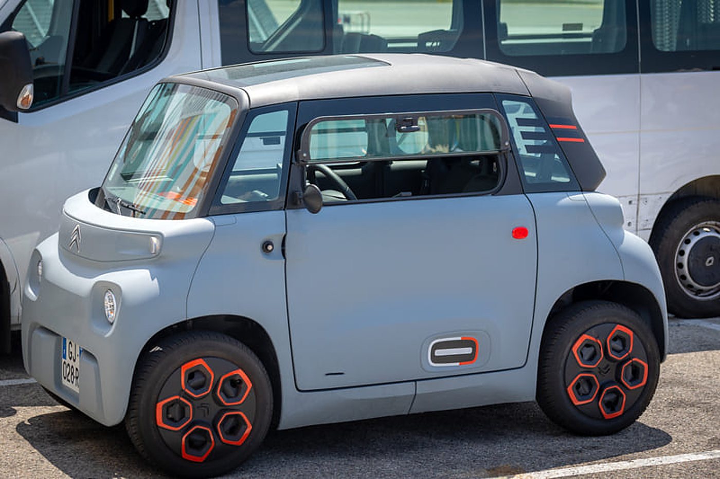 Une petite voiture électrique grise aux jantes décorées d'orange est garée sur un parking devant des camionnettes blanches.