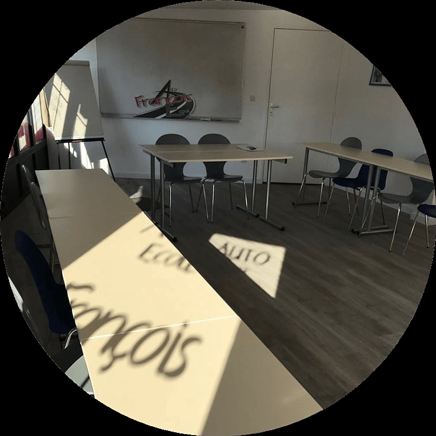 Salle de formation lumineuse d'une auto-école avec des tables alignées, des chaises, un tableau blanc portant un logo et des ombres de lettres sur les tables.