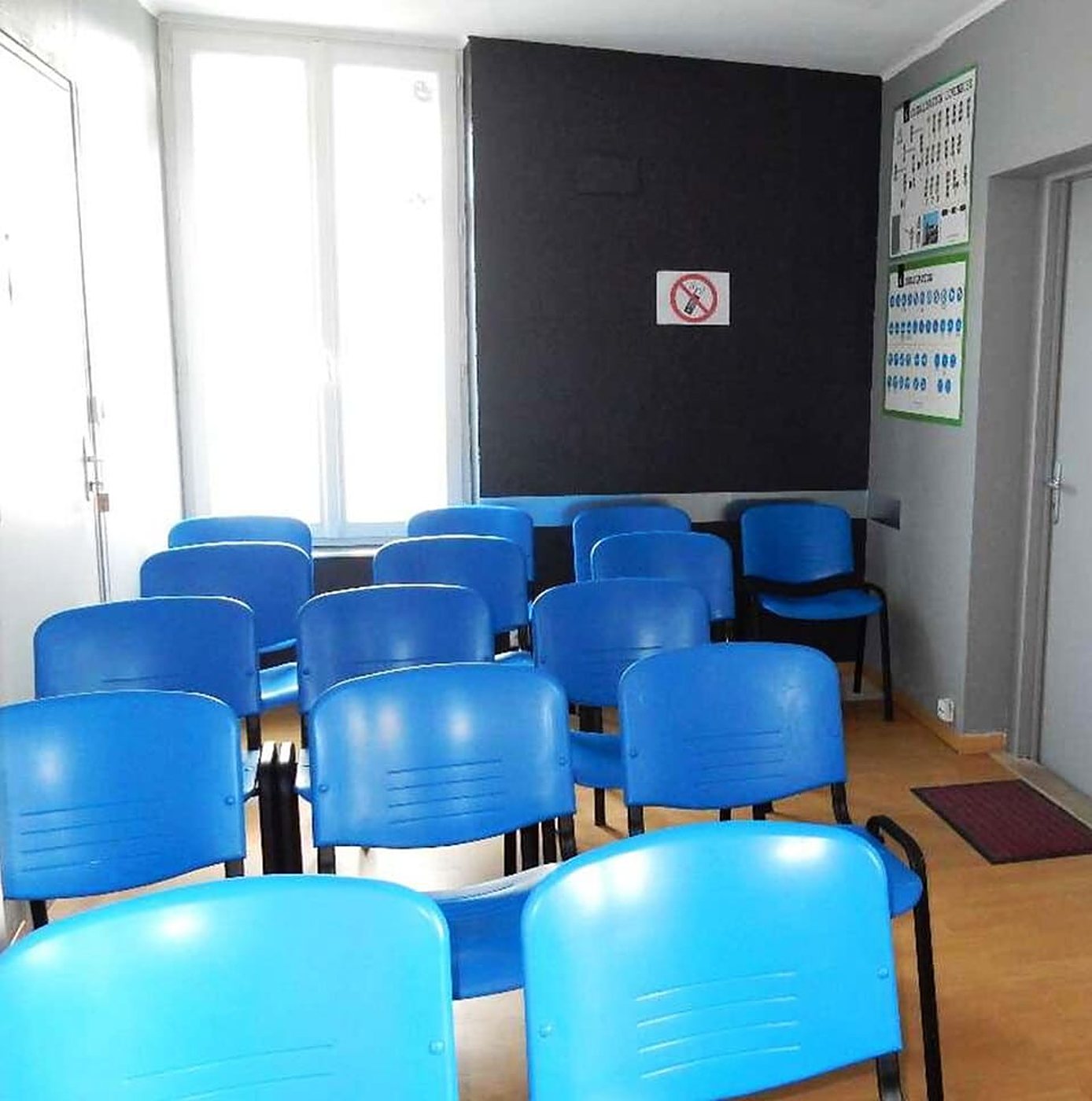 Une petite salle de formation lumineuse avec des rangées de chaises bleues face à un mur noir, des affiches au mur et un panneau d'interdiction de téléphoner.