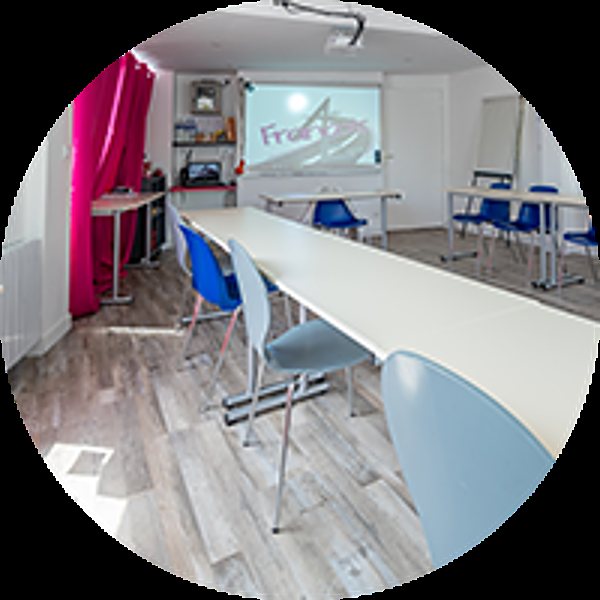 Salle de formation avec de longues tables blanches, des chaises bleues, un rideau rose et un écran de projection au fond.