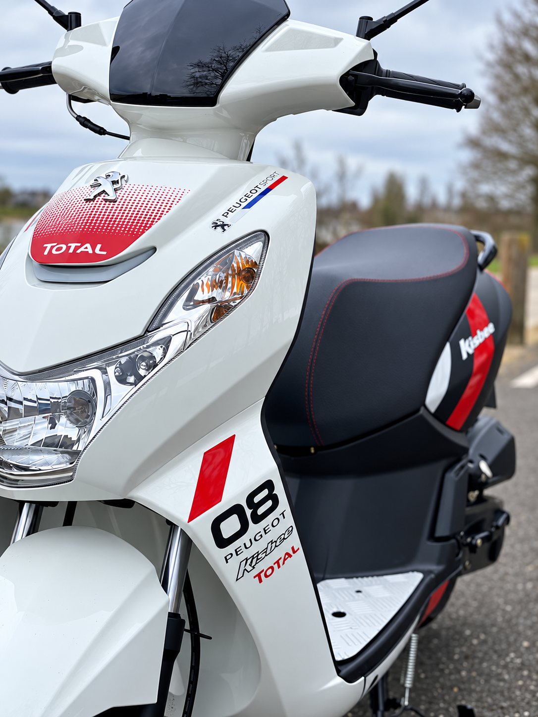 Vue de face d'un scooter Peugeot Kisbee blanc, orné d'autocollants Total et du numéro 08, garé sur une route à l'extérieur.