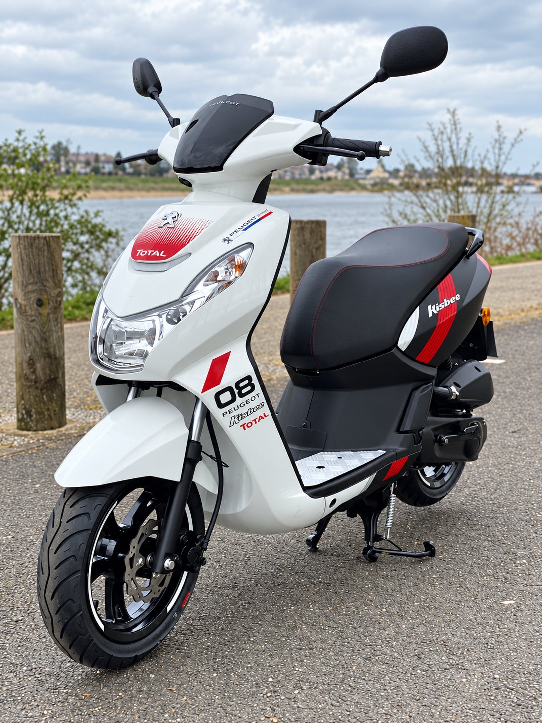 Un scooter Peugeot Kisbee blanc et noir, orné de décors rouges Total, est garé sur une route au bord d'un plan d'eau.