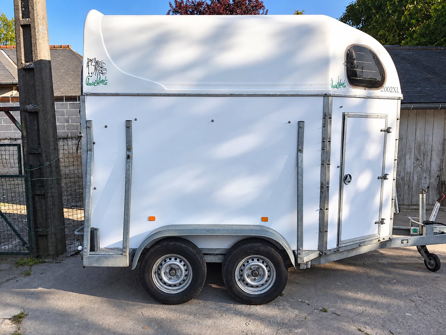 Un van blanc pour le transport de chevaux, équipé de deux roues, est stationné dans une cour devant un grillage et des bâtiments.