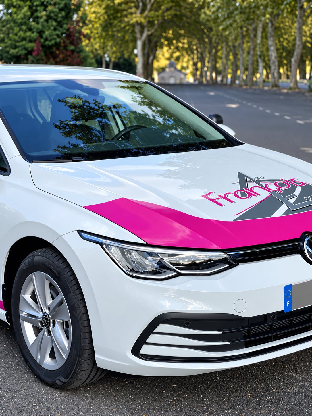 Avant d'une voiture blanche aux bandes roses, avec le logo François sur le capot, garée sur une route bordée d'arbres.