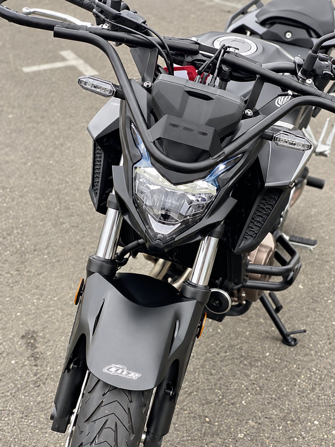 Une moto roadster noire vue de face, avec son phare et son guidon, est garée sur un parking en bitume, avec d'autres motos en arrière-plan.